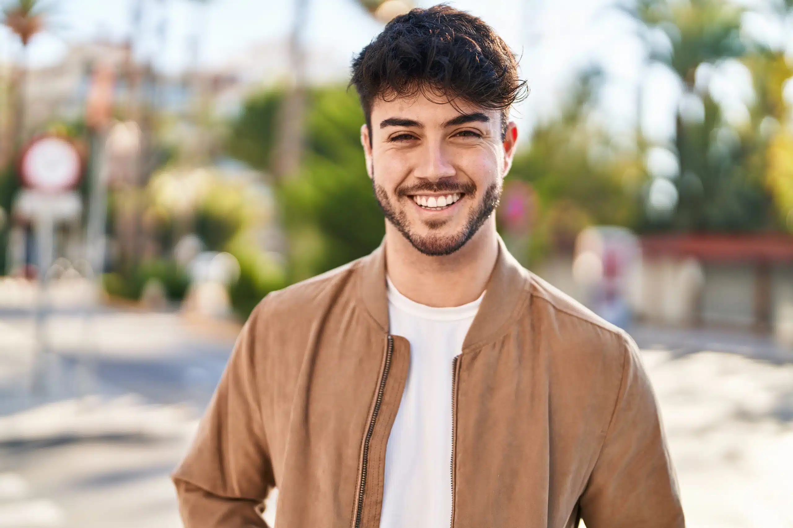 Smiling young man with dark hair standing outdoors in a casual brown jacket, representing confidence and a healthy appearance.