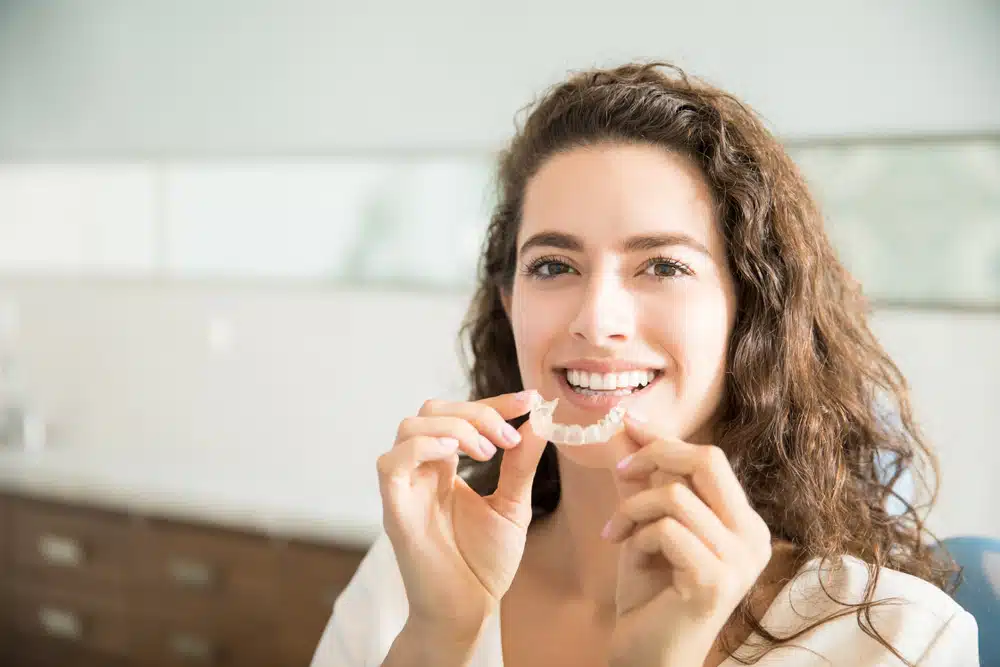 Woman smiling while holding a clear Invisalign aligner for discreet orthodontic treatment